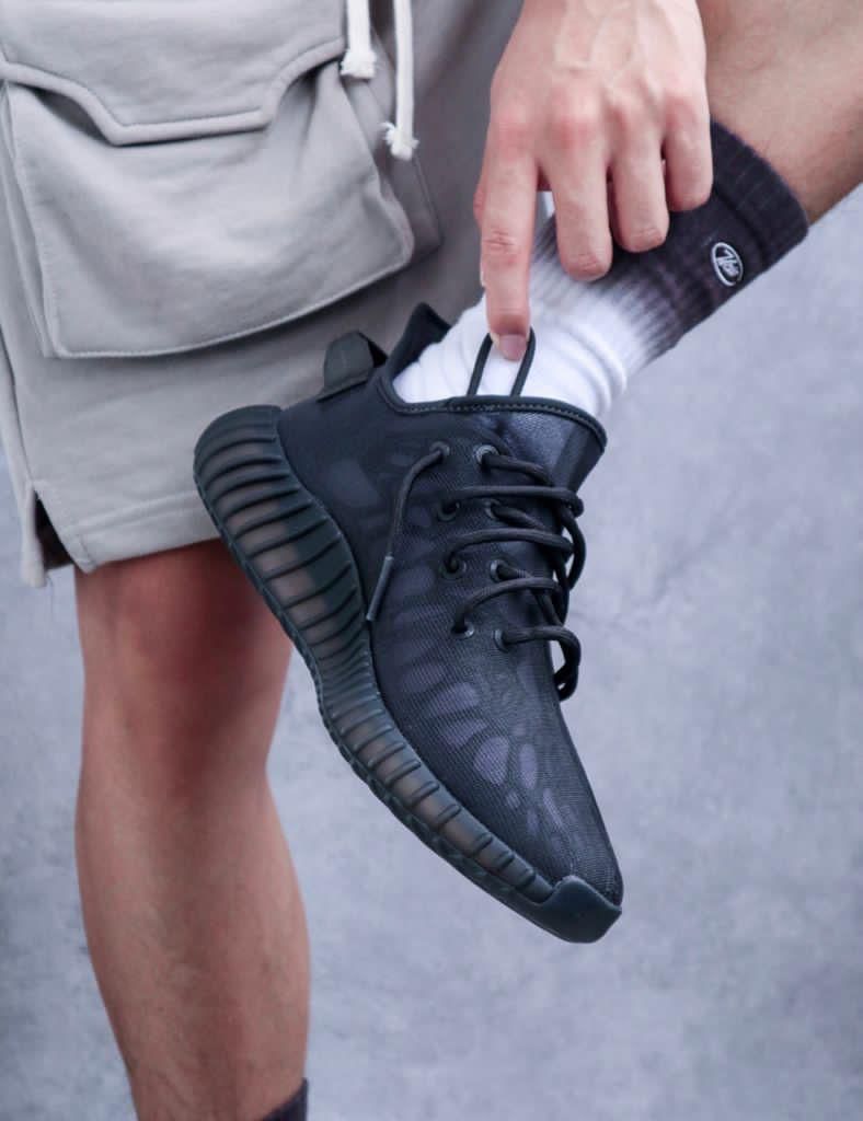 dffef36c-2a85-4bad-9087-e0ba836c07ae_f7fd0796-e162-4e4b-89ae-7d1fa80a06c5 | AE JERSEY Person wearing a black athletic shoe and white sock, lifting their foot to adjust the laces against a beige short.