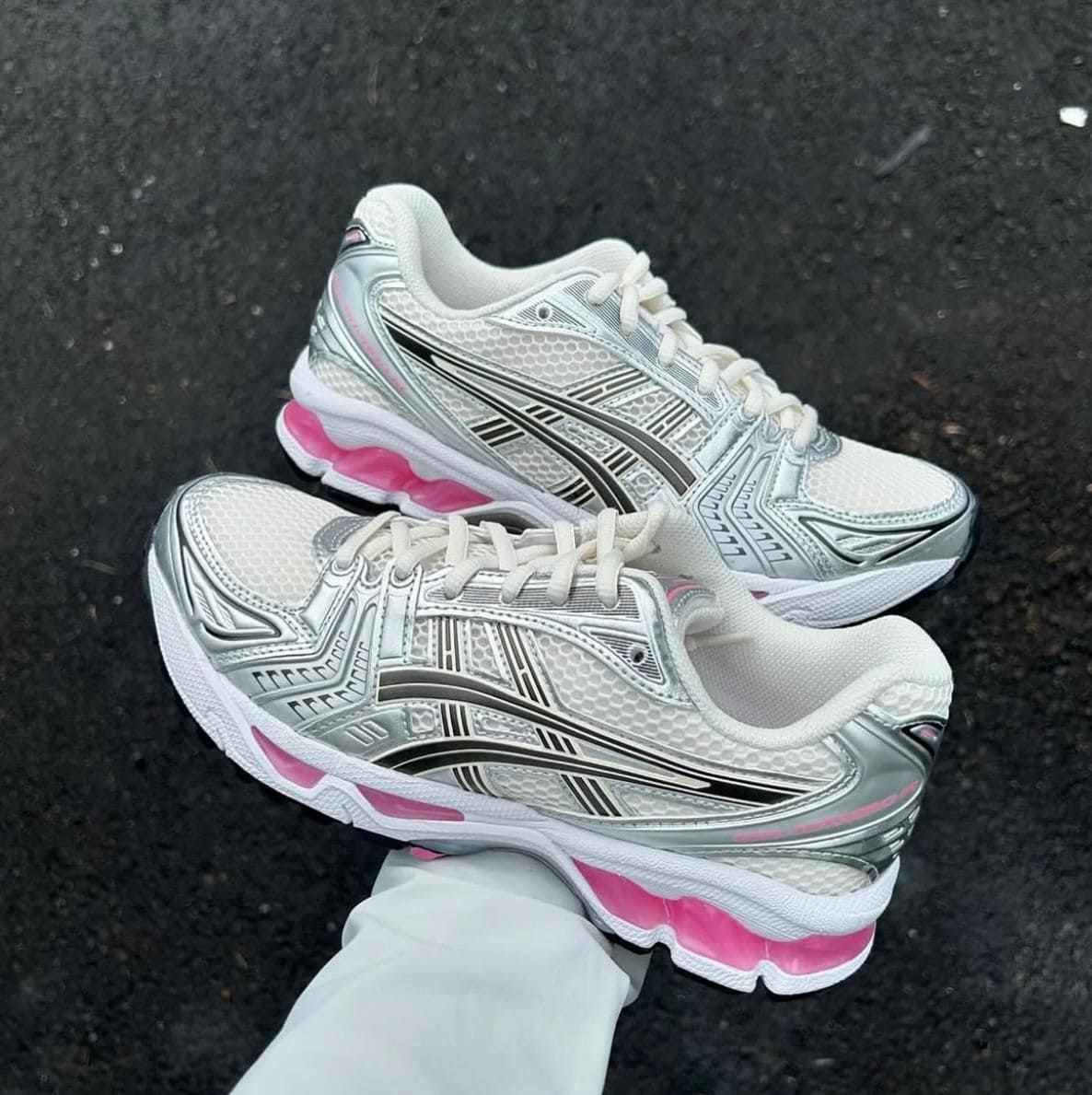 e0909508-6cc4-42df-a209-13c88a42025c | AE JERSEY Pair of white and silver running shoes with pink accents resting on dark pavement