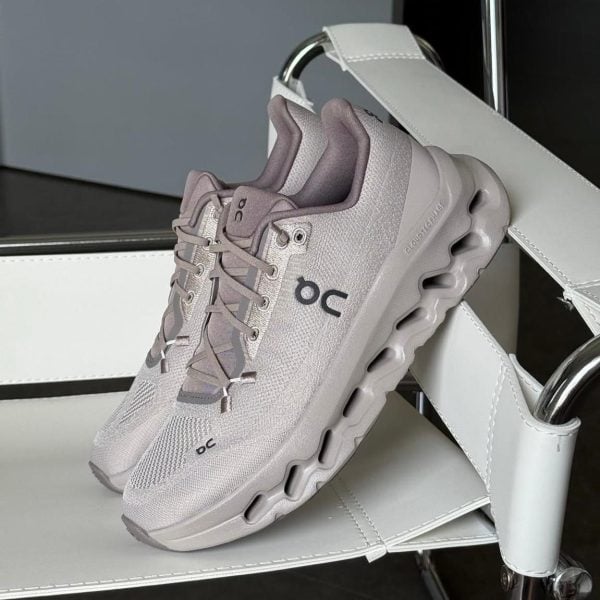 Light gray On running sneaker with a cloud-like sole resting on a white display stand in a showroom setting.