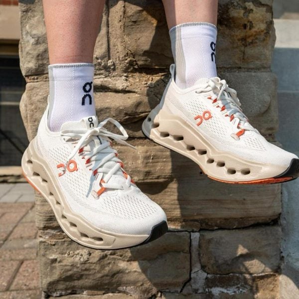 White athletic sneakers with orange accents and chunky beige sole, worn with white crew socks on stone steps, casual outdoor scene.