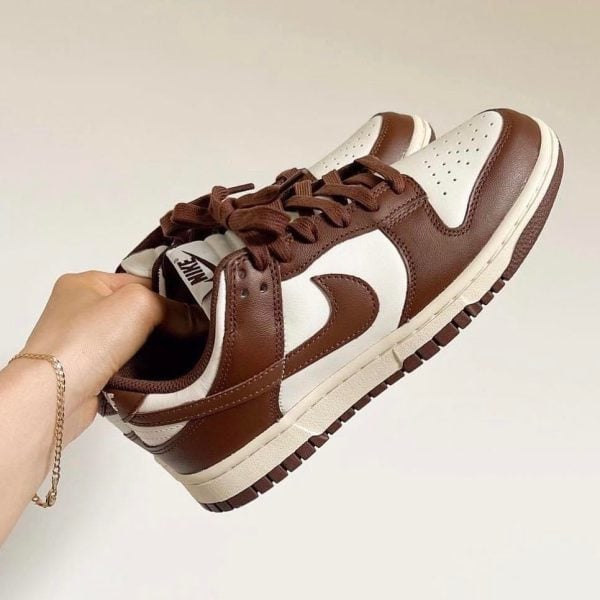 Brown and white Nike sneakers held in a hand, showing the side view with perforated toe and white swoosh logo on brown panels.