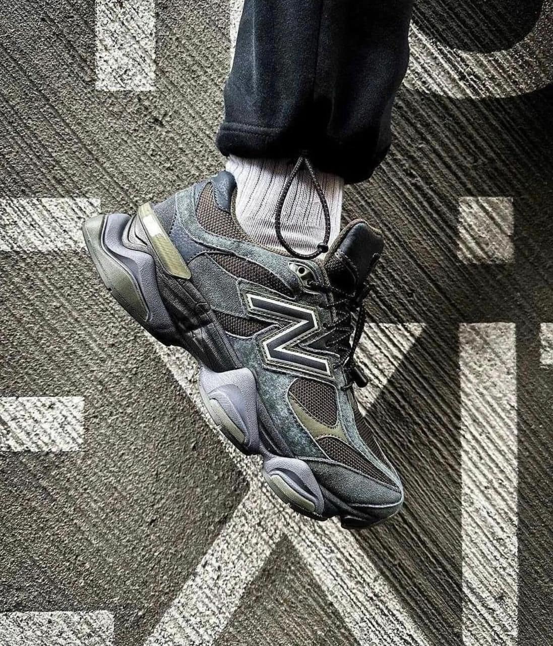 e8b95b1a-ab1e-4c5c-9fda-93eb9564c313 | AE JERSEY Close-up of a dark gray New Balance-style sneaker with a bold side logo, laces tied, worn with a light gray sock, resting on a textured, patterned carpet.