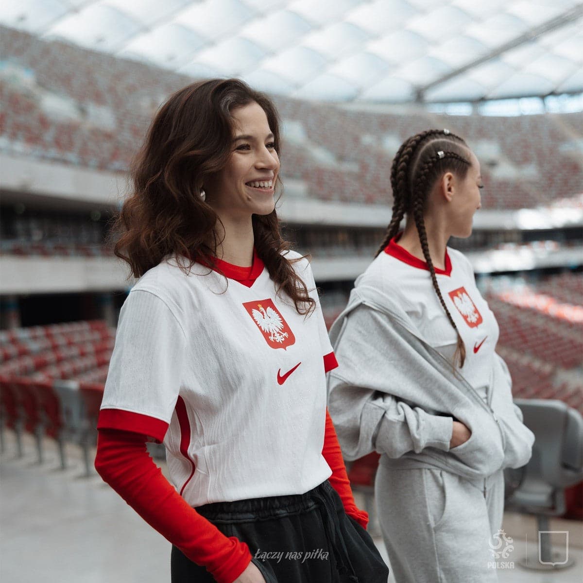 eJCGc4HHxhcUUll | AE JERSEY Two smiling young women in white Poland jerseys stand side by side in a stadium, one with red sleeves visible beneath.