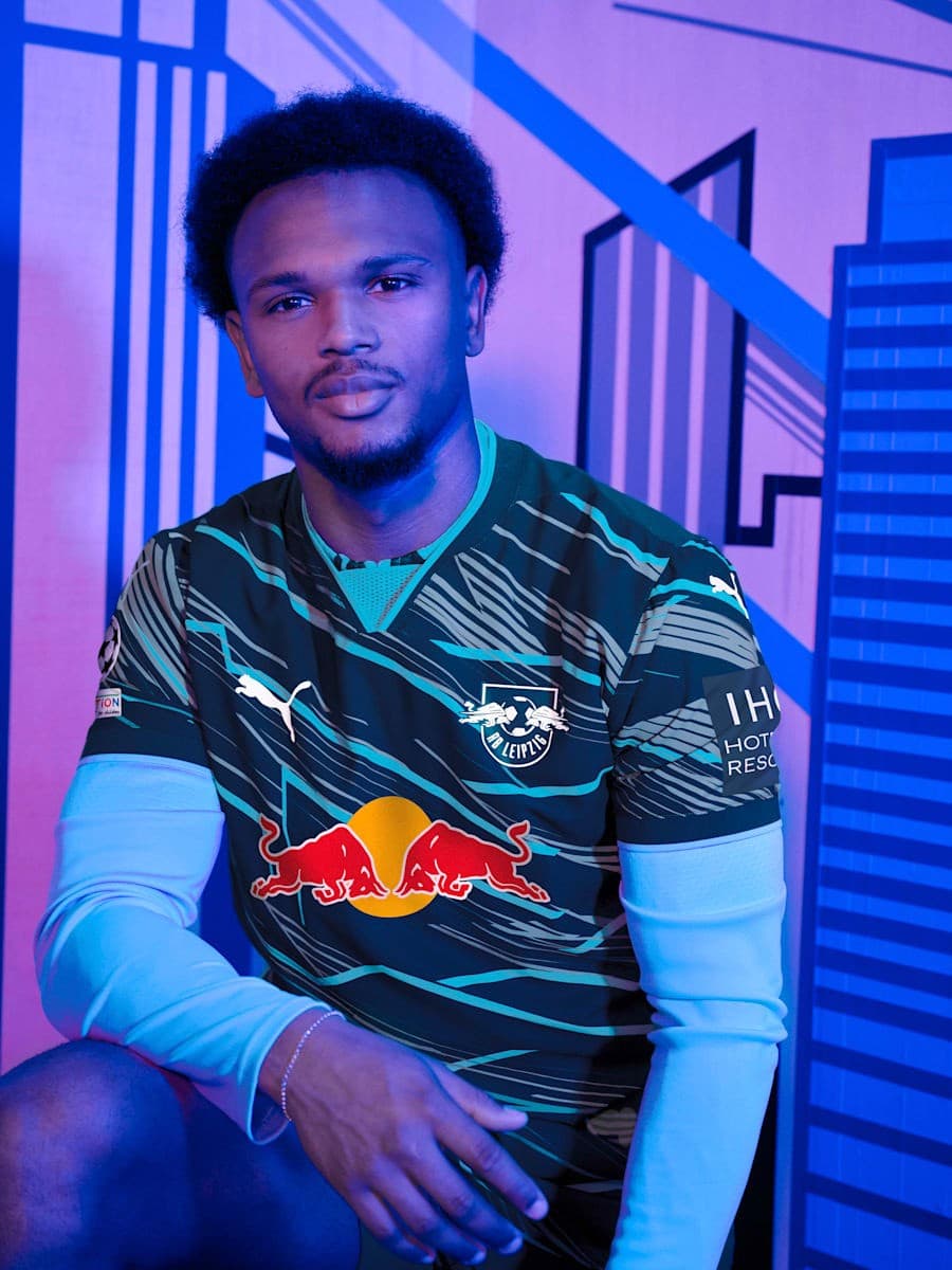 eZ7ewnAE4NkyyL6 | AE JERSEY Young man seated in a sports jersey with a Red Bull logo, posing casually in a blue-lit backdrop with geometric patterns.