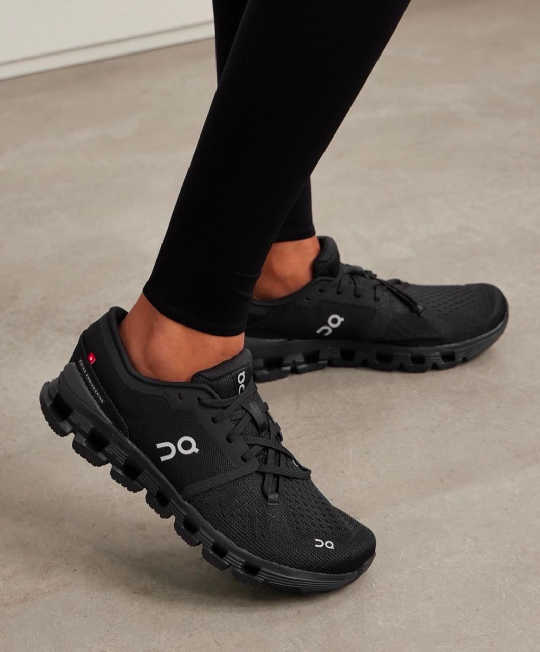 f2456ba2-2709-4361-8435-d5d5ea96bafe | AE JERSEY Person wearing black athletic shoes and black leggings, standing on a light floor, showing the On logo on the sides.