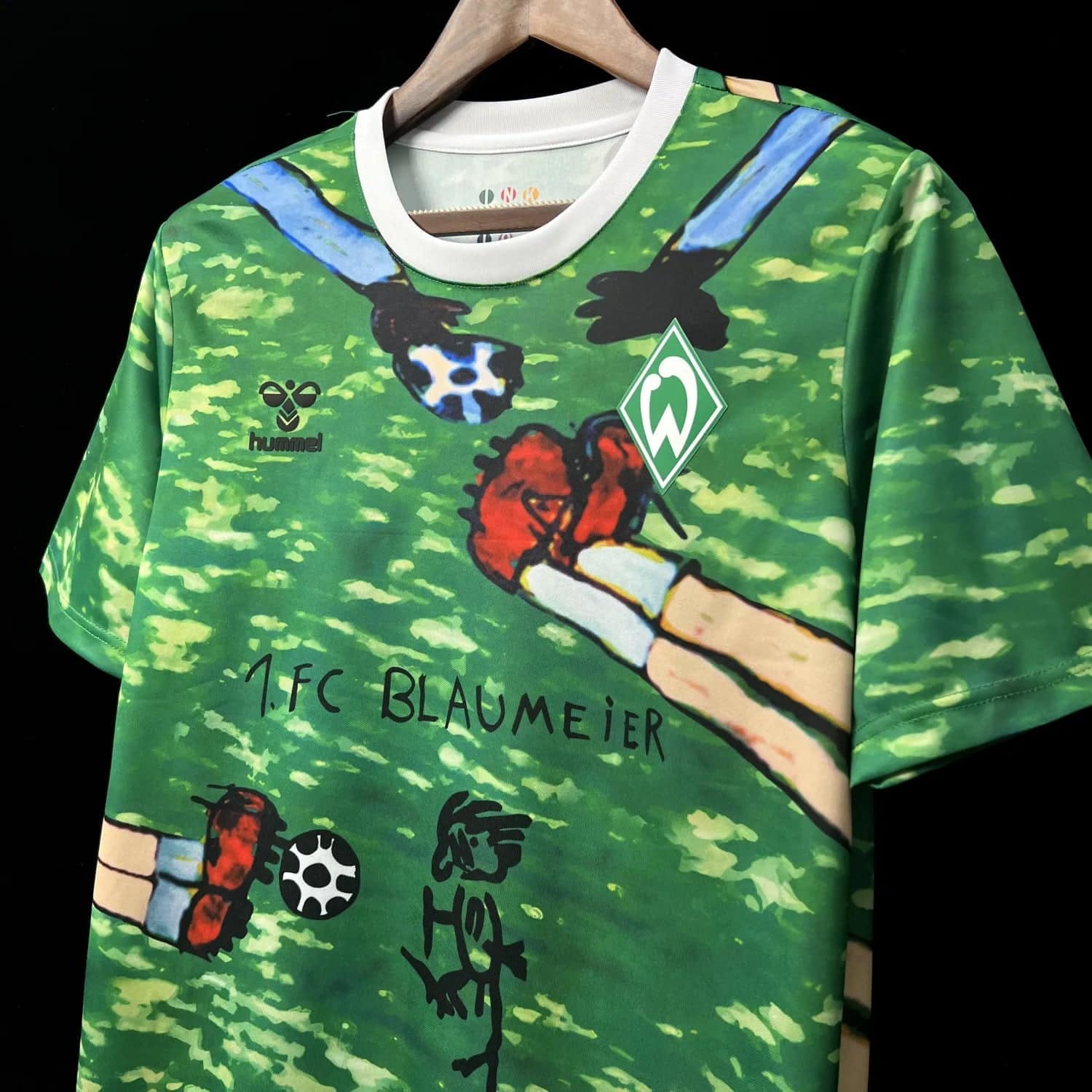f2547a8d0d4c9e86 | AE JERSEY Green sports jersey on a hanger with a cartoon hand graphic, Werder Bremen crest, and Hummel logo; handwritten '1.FC BLAUMEIER' text visible.