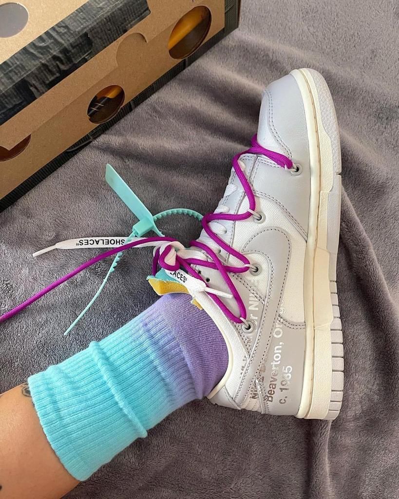 f4d0bfce-af77-4eeb-b612-31cb19fda118_9d9ce0df-d3b4-43ed-9ccd-14143502c6e9 | AE JERSEY White high-top sneaker with purple laces on a gray blanket, turquoise socks showing, and a cardboard shoebox in the background.