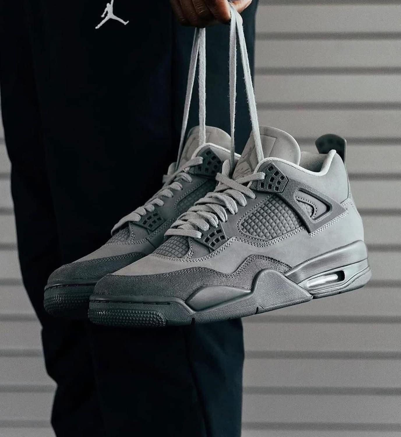 f58e7aff-18c9-4cc9-8c3f-0531646c732d | AE JERSEY Gray athletic sneakers are being held by their laces in front of a dark outfit and a gray siding background.