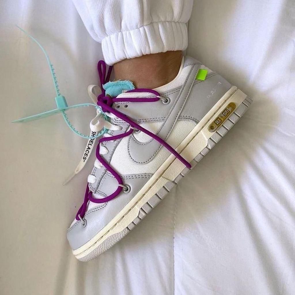 f7df711c-5bcb-4d6f-a0d1-83b15b488c53_5cabee29-e3ad-435a-ad99-7677b371cce3 | AE JERSEY White low-top sneaker with bright purple laces and teal tag, resting on a white bedsheet with a cuffed pant leg visible.