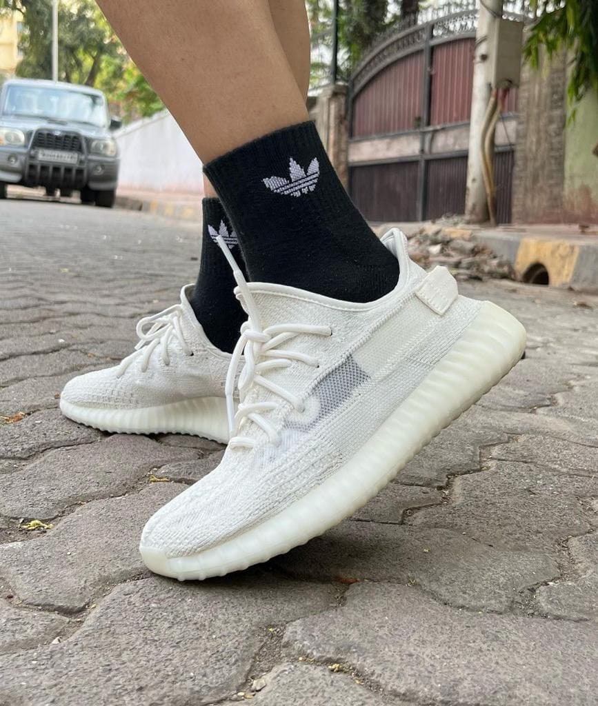 f8746cc7-8bf6-4a62-9fdb-cd7d199bba95_0b3589e7-fdcd-4255-b82d-75d475bf8c7b | AE JERSEY White knit sneakers with laces being worn with black Adidas socks on a cobblestone street, mid-step in an urban setting.