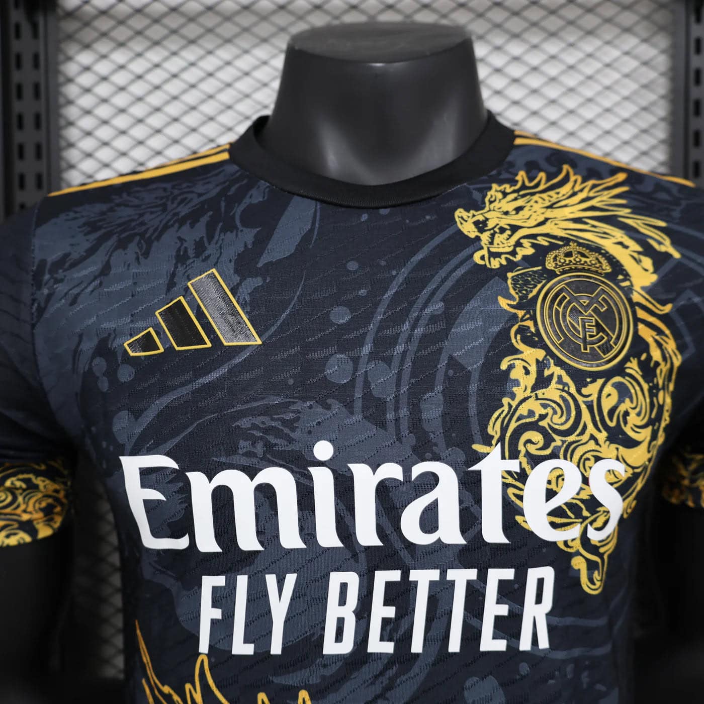 fdbf06cb | AE JERSEY Close-up of a Real Madrid soccer jersey with a black‑gold pattern, crest on the right chest, and Emirates Fly Better slogan visible.