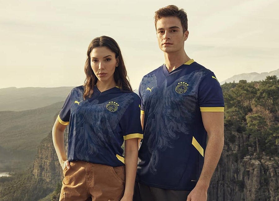 fenerbahce_24_25_puma_fourth_kit | AE JERSEY Two models stand outdoors in navy blue and gold soccer jerseys with a canyon backdrop.