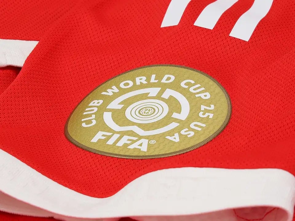 filters_no_upscale__format_webp__quality_50 | AE JERSEY Gold circular patch with FIFA logo on a red sports jersey, surrounded by white trim lines and fabric texture.