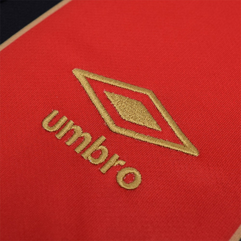 g7kJQvN0i0hobWT | AE JERSEY Close-up of red fabric with gold embroidery of the Umbro logo and brand name.