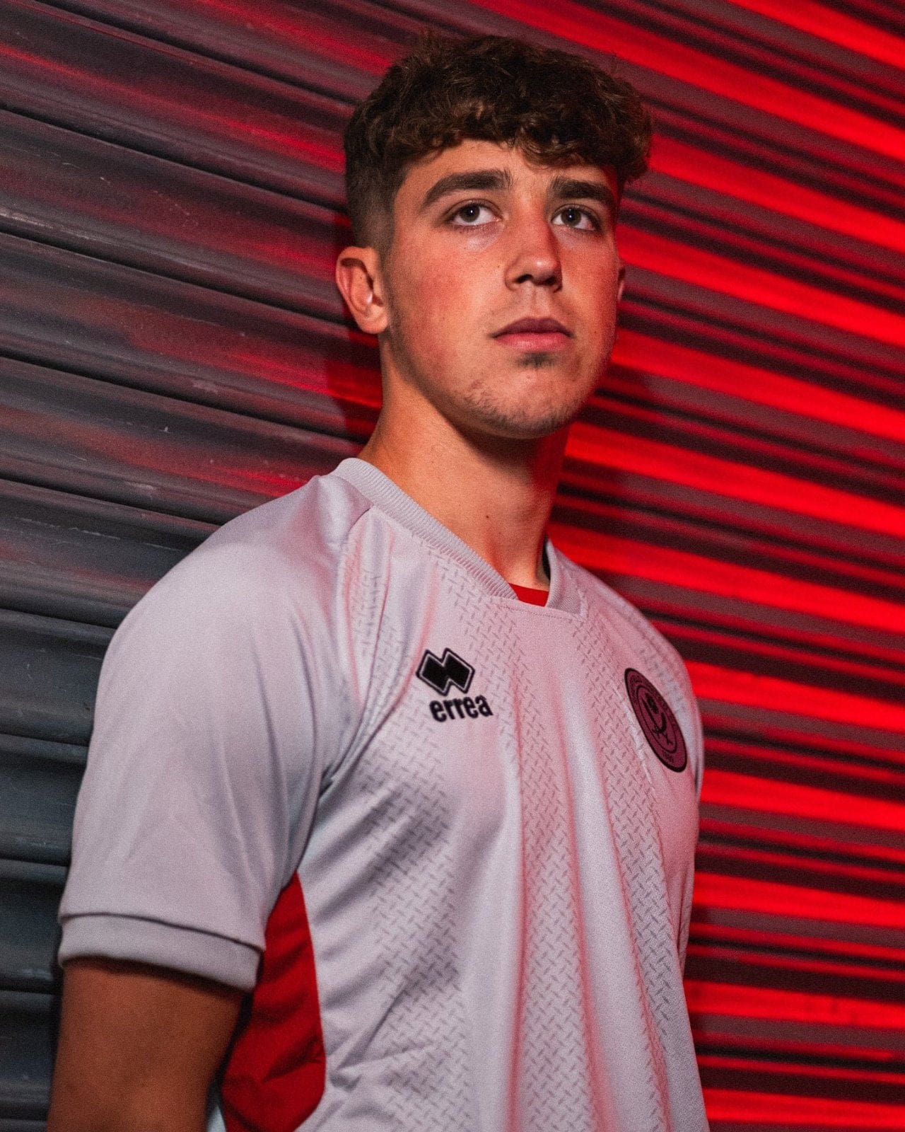 gZGDEAqUutD92Q9_3e2cf362-1e74-4b4d-8e96-324ac093c336 | AE JERSEY Portrait of a young man in a white team jersey with red side panels, standing against a red-lit striped background.