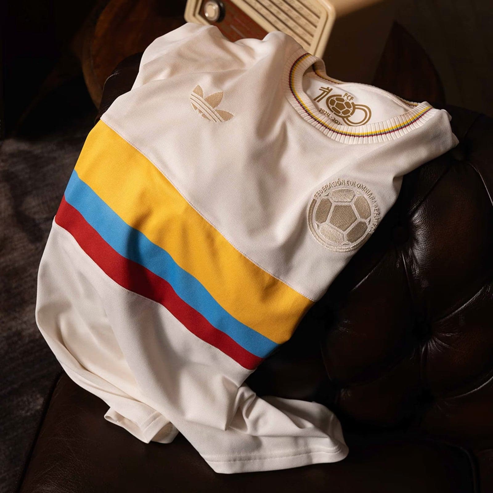gylaggfgziPYWk4 | AE JERSEY White Colombia soccer jersey with yellow, blue, and red stripes lying on a dark brown couch; Adidas logo and national crest visible nearby.