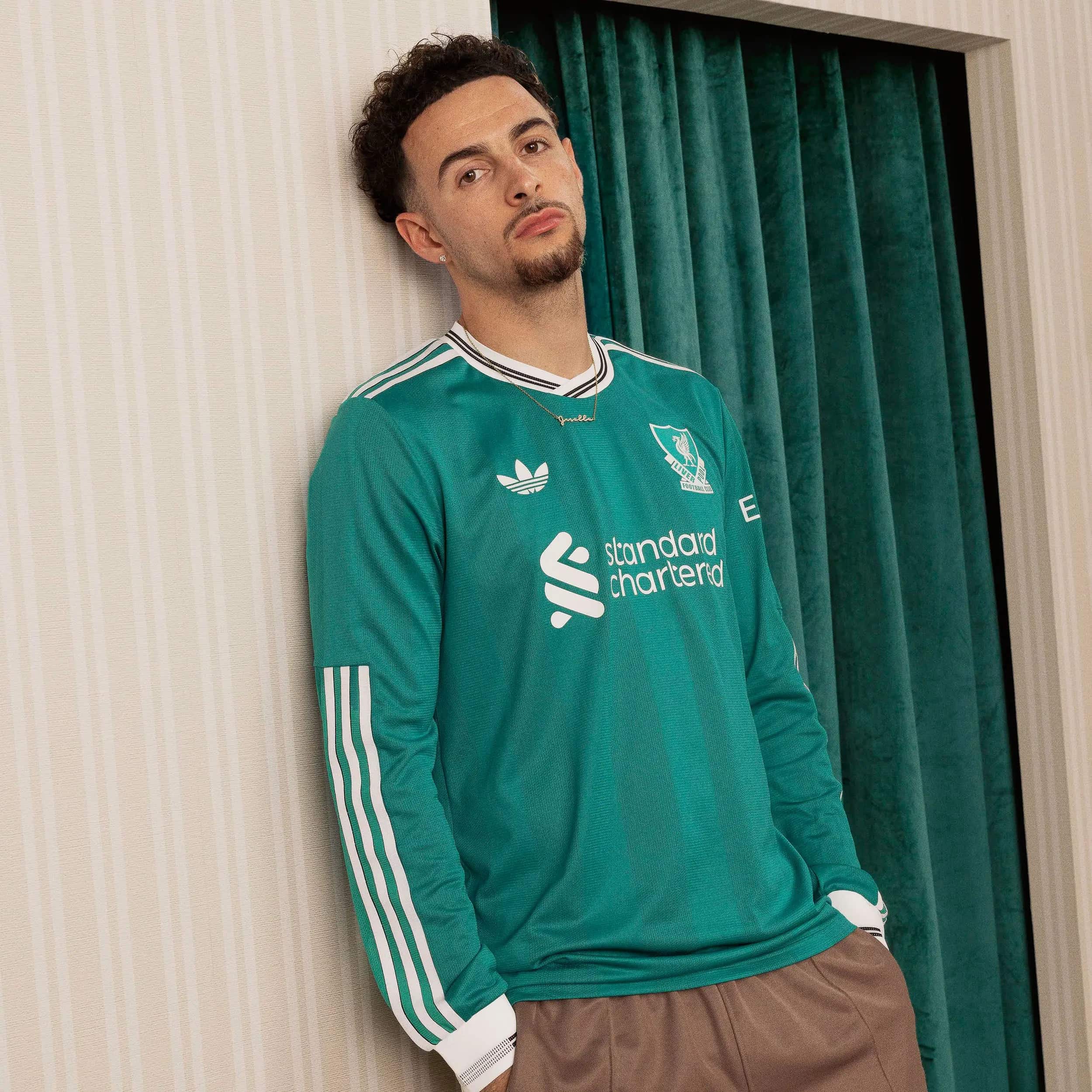 h24536-fw25_liverpoolfc_toolkit_third_adidas_jones_0683-1352257_1 | AE JERSEY Young man leaning against a beige striped wall in a teal football jersey with sponsor logos visible on the front.