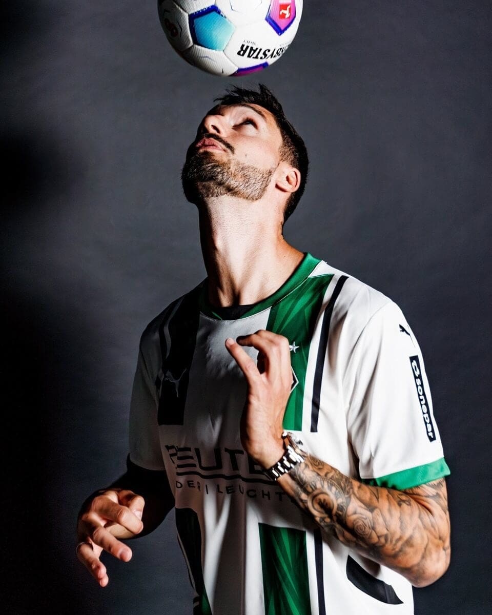 iN100zKGt0sG6jr | AE JERSEY Man in white and green soccer jersey balances a ball above his head, looking up at it in a studio.