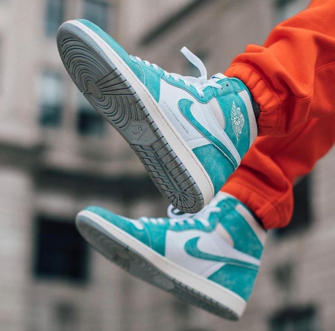 image_7dad14b6-dc4b-4ef3-9817-c844e1551b98 | AE JERSEY Teal and white high-top sneakers in mid-air against a blurred urban backdrop, orange pants visible at the edges.