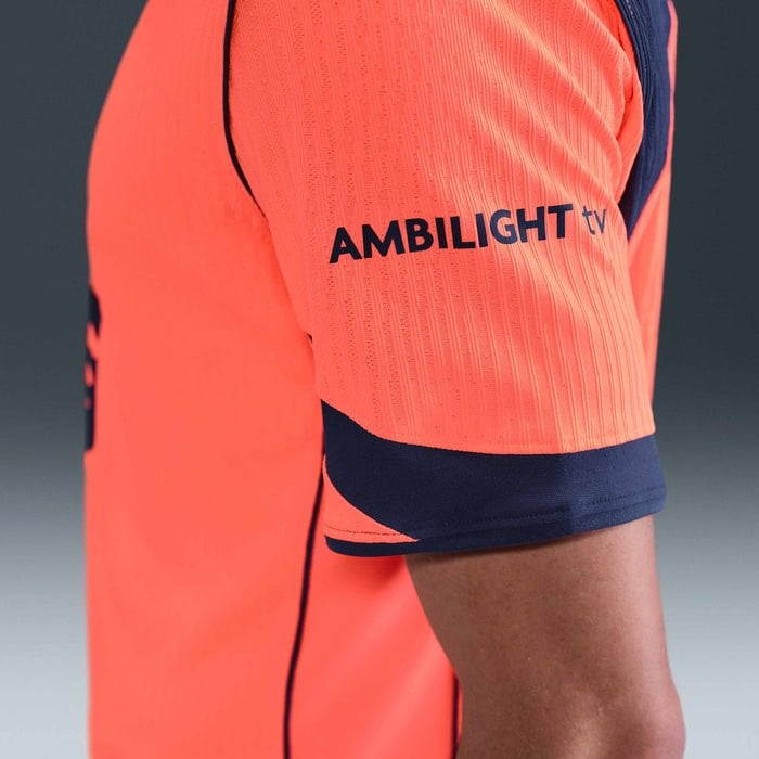 kd | AE JERSEY Close-up of a bright orange sports jersey sleeve with navy trim and the text 'AMBILIGHT TV' on the upper arm.