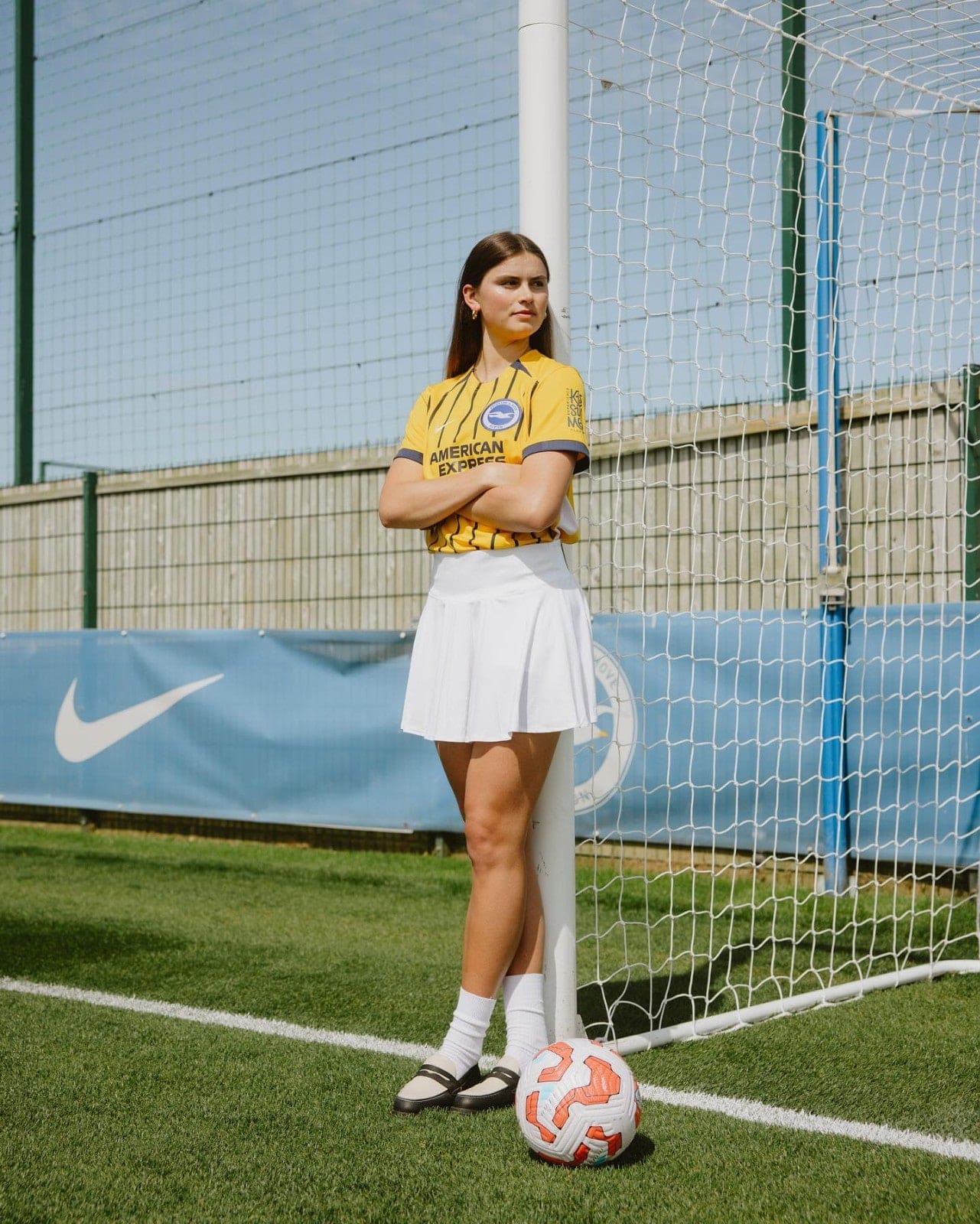 lYJAstBUgBZMQyf | AE JERSEY Young woman in a yellow soccer jersey and white skirt stands with arms crossed beside a goal post, a soccer ball at her feet on a sunny field.