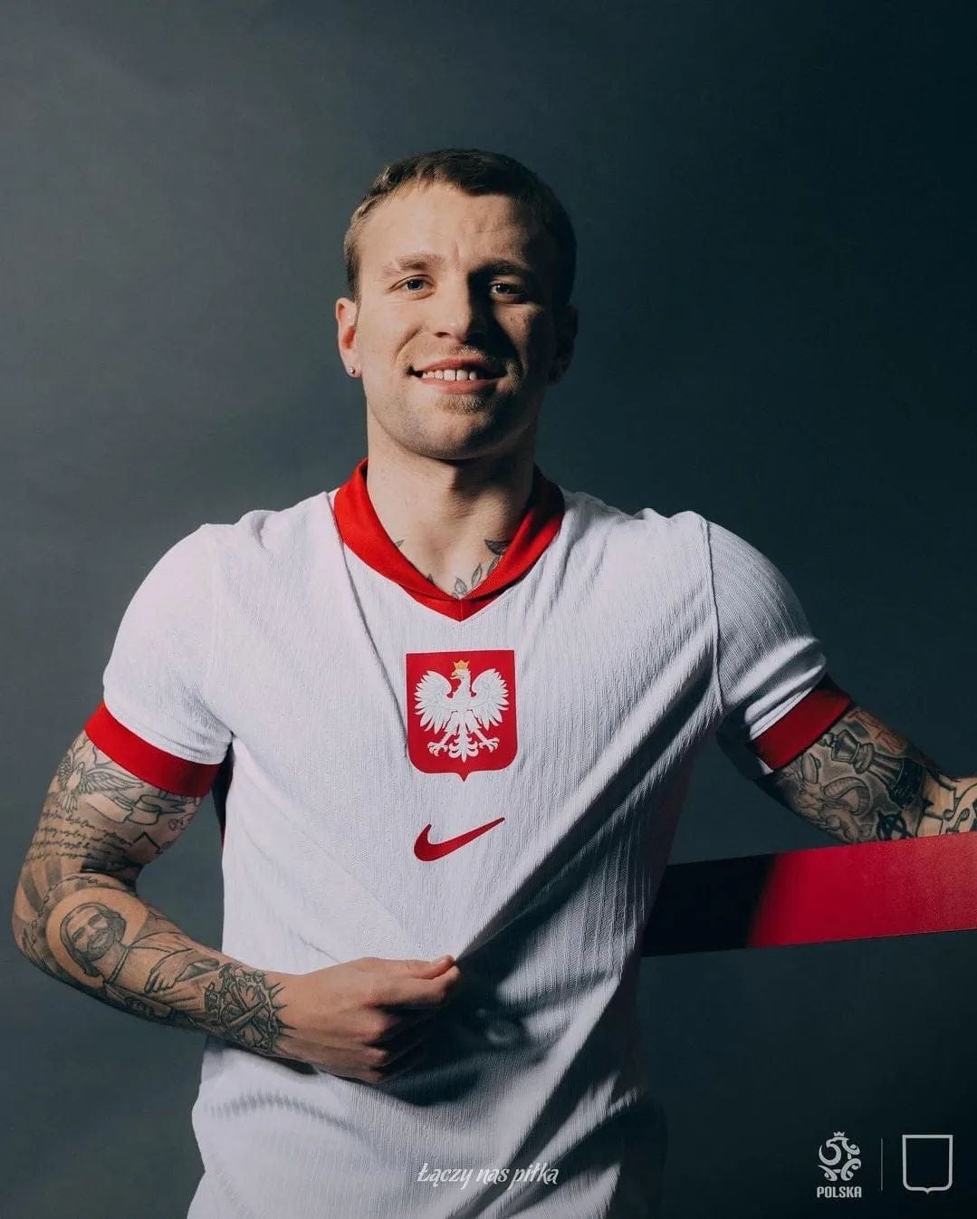 m6eAyBzCOghPdkI | AE JERSEY Polish footballer wearing a white jersey with red eagle crest and Nike logo, smiling with tattoos on both arms against a dark background.