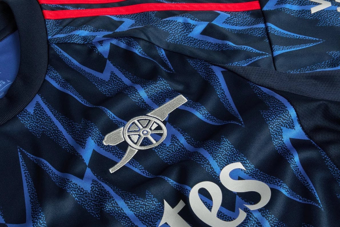 mji9511_d1 | AE JERSEY Close-up of a navy blue-patterned football jersey featuring the silver Arsenal cannon crest and partial sponsor text.