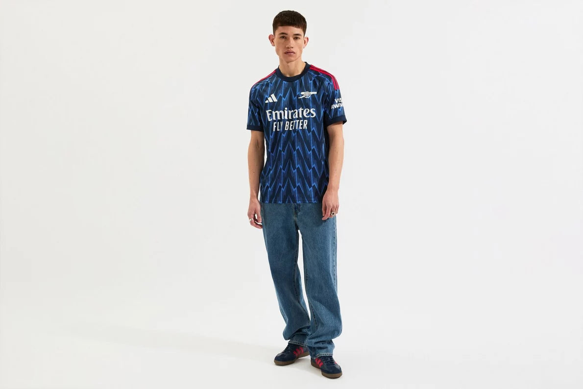 mji9511_f2 | AE JERSEY Male model wearing a blue zigzag-pattern Arsenal football jersey with 'Emirates Fly Better', jeans, and dark sneakers.