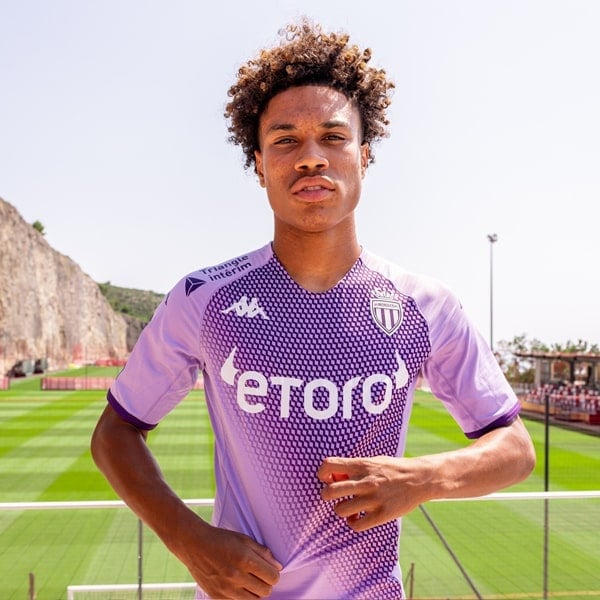 monaco-tab-min_128e5f43-d88f-43fc-9505-f612706b36f3 | AE JERSEY Soccer player in a purple kit standing on a bright green field, posing confidently toward the camera with hands on hips.