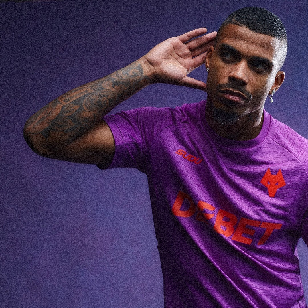 n16MKVhOxXi5QYG | AE JERSEY Man in a purple sports jersey posing with hand near his ear, looking to the side; the shirt features a red DEBET logo and a fox emblem on the chest.