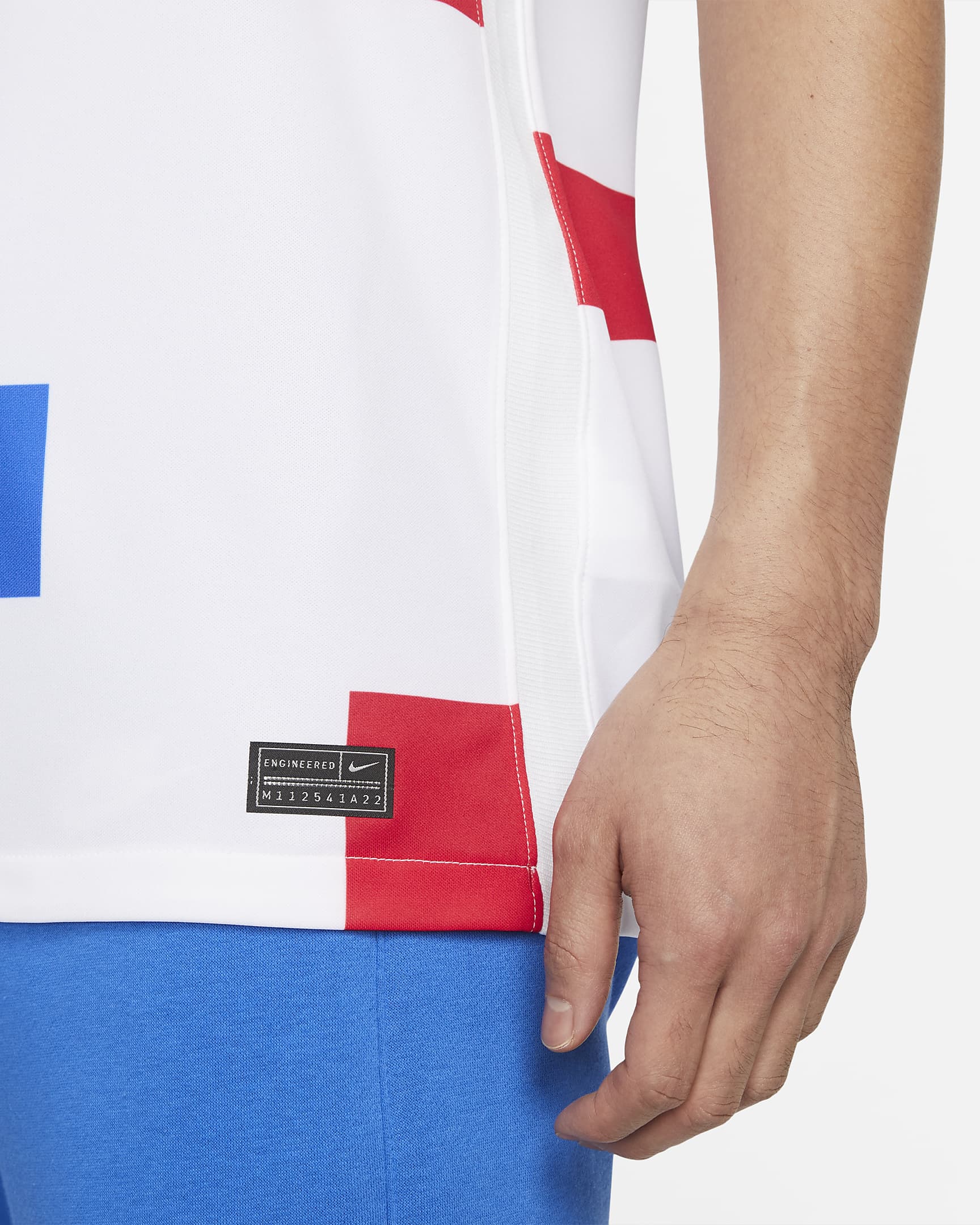 netherlands-2022-stadium-away-football-shirt-XRm5hx_37e99650-d934-453c-9841-116d3a8fd423 | AE JERSEY Close-up of a person wearing a white jersey with red panels and blue shorts, focusing on the torso area and arm at the side.