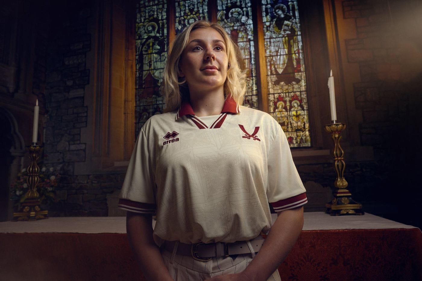 oo3RB5CblNufCNn | AE JERSEY Young woman in a cream polo with burgundy trim stands at an altar in a church with stained glass behind her.