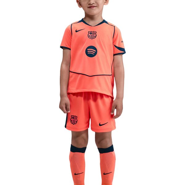 Young boy in a bright orange FC Barcelona soccer kit, standing and smiling with neon yellow cleats on a white background.