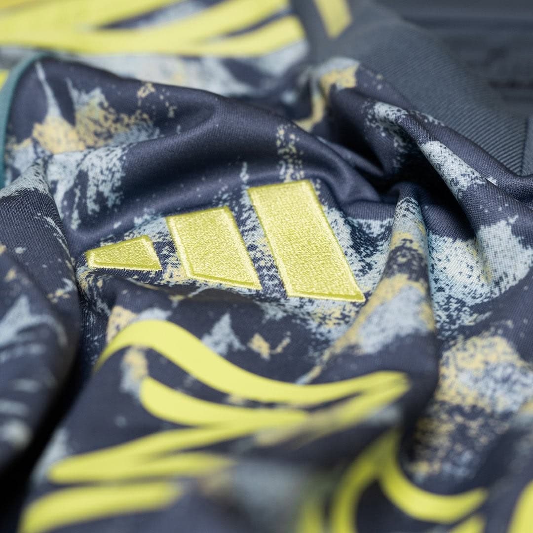 pAYMdutXuXkN8E6 | AE JERSEY Close-up of dark navy fabric with a bright yellow stitched three-stripe emblem on a camo-like pattern.