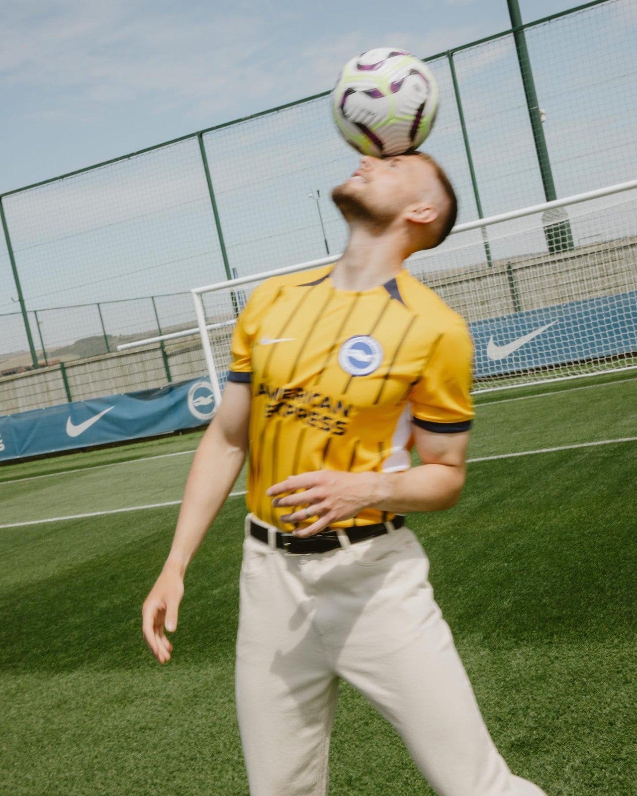qwqYOIkPVvyKzMK | AE JERSEY Soccer player in a bright yellow striped jersey heads a soccer ball on a fenced practice field.