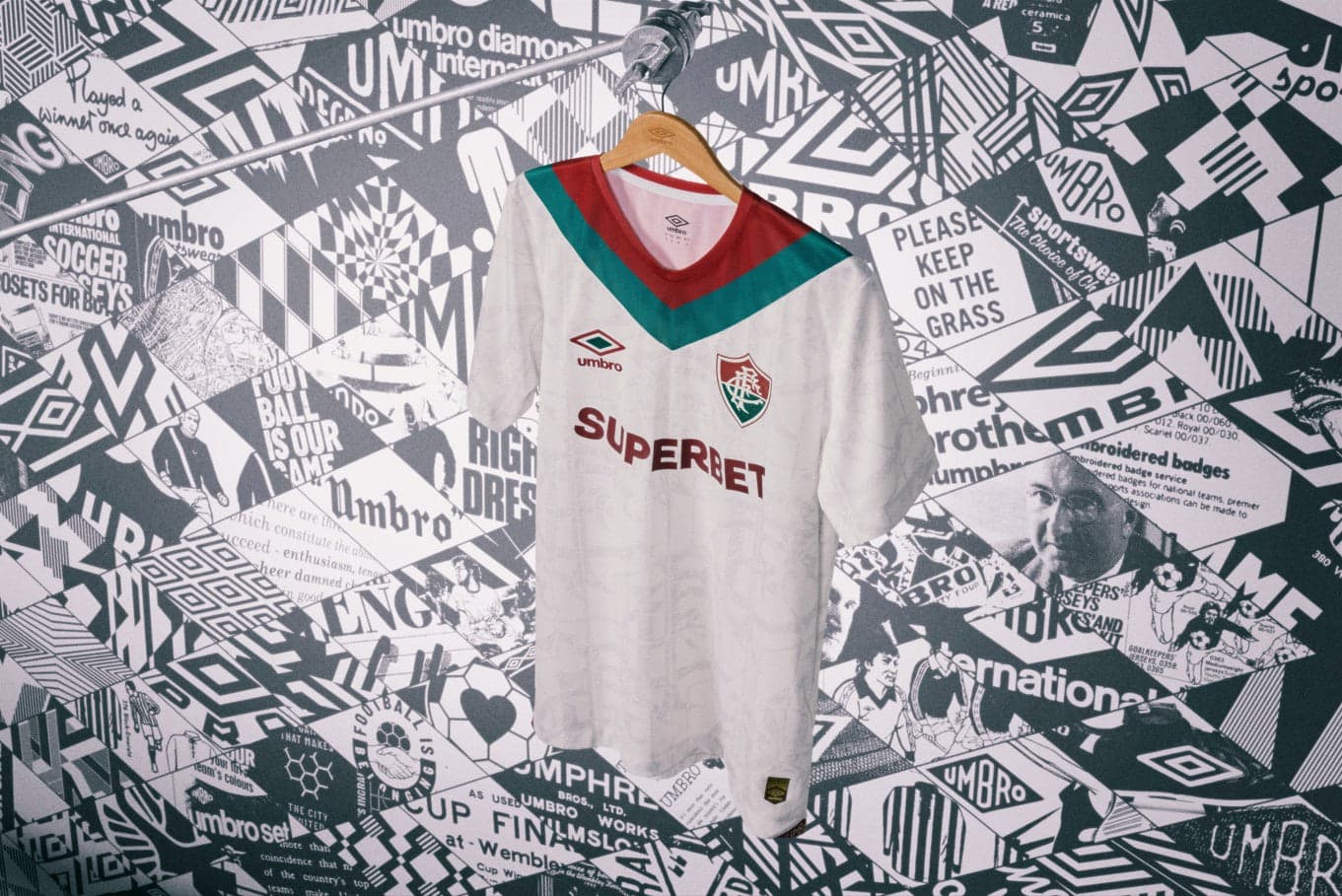 qyf8tIMkWz7jVGq | AE JERSEY White football jersey on a hanger in front of a collage wall; features a green and red V-neck, Umbro logo, club crest, and the sponsor text 'SUPERBET'