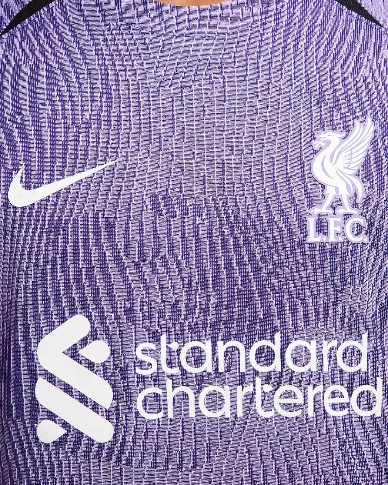 rn-image_picker_lib_temp_0c24d7af-4250-431b-9757-f13a50c77087 | AE JERSEY Close-up of a purple Liverpool FC jersey featuring the white Nike swoosh and the club crest with sponsor text nearby.