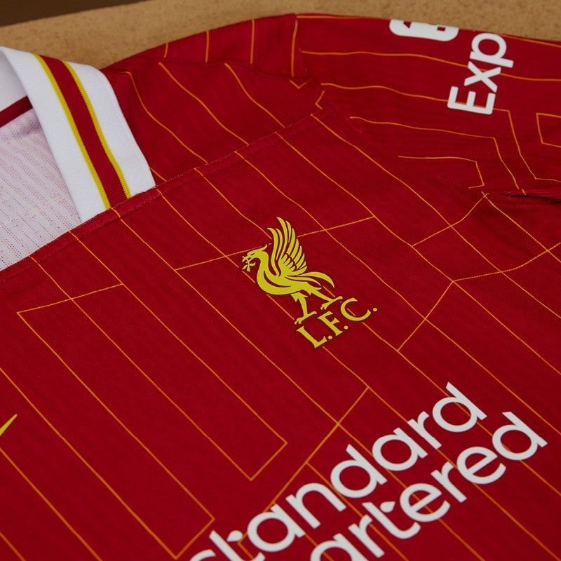 rn-image_picker_lib_temp_296fa7a6-0c70-483b-9b71-c06ef3381568 | AE JERSEY Red Liverpool FC jersey with yellow crest and geometric orange lines, white sponsor text visible at the bottom edge, collar detail nearby.