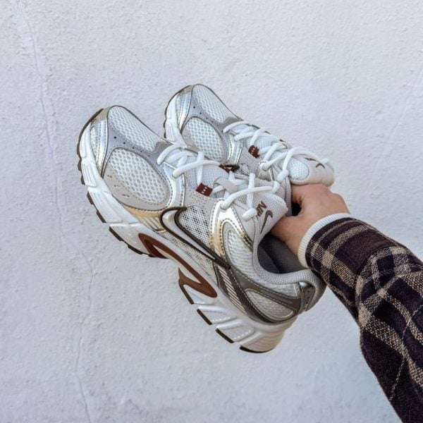 A person holding a pair of white and silver running shoes against a light wall backdrop.