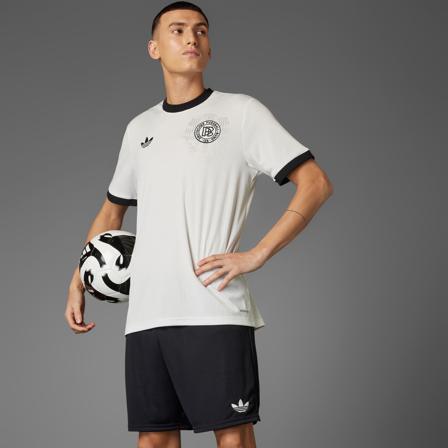 rn-image_picker_lib_temp_3d50a883-16c2-40c9-8c49-2d44526dc3c4 | AE JERSEY Athlete in a white soccer jersey holding a black-and-white soccer ball against a gray backdrop.