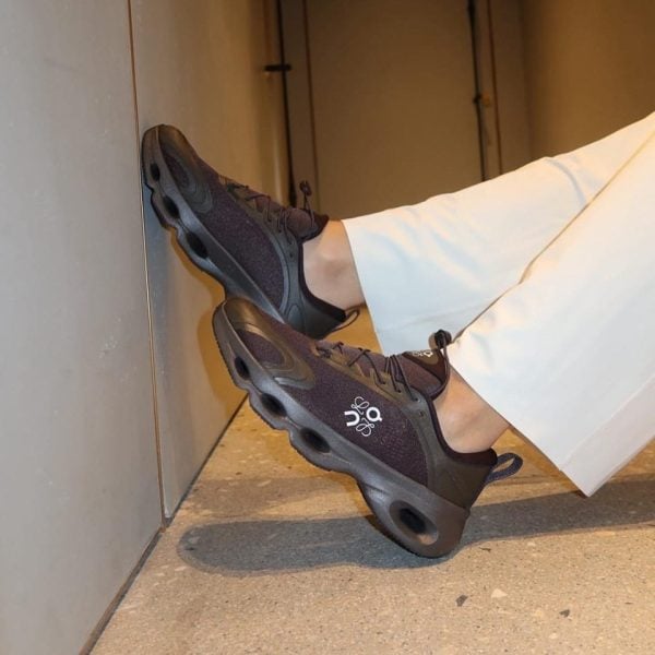 Person wearing dark purple athletic sneakers with a chunky black sole, feet propped against a wall in white pants.