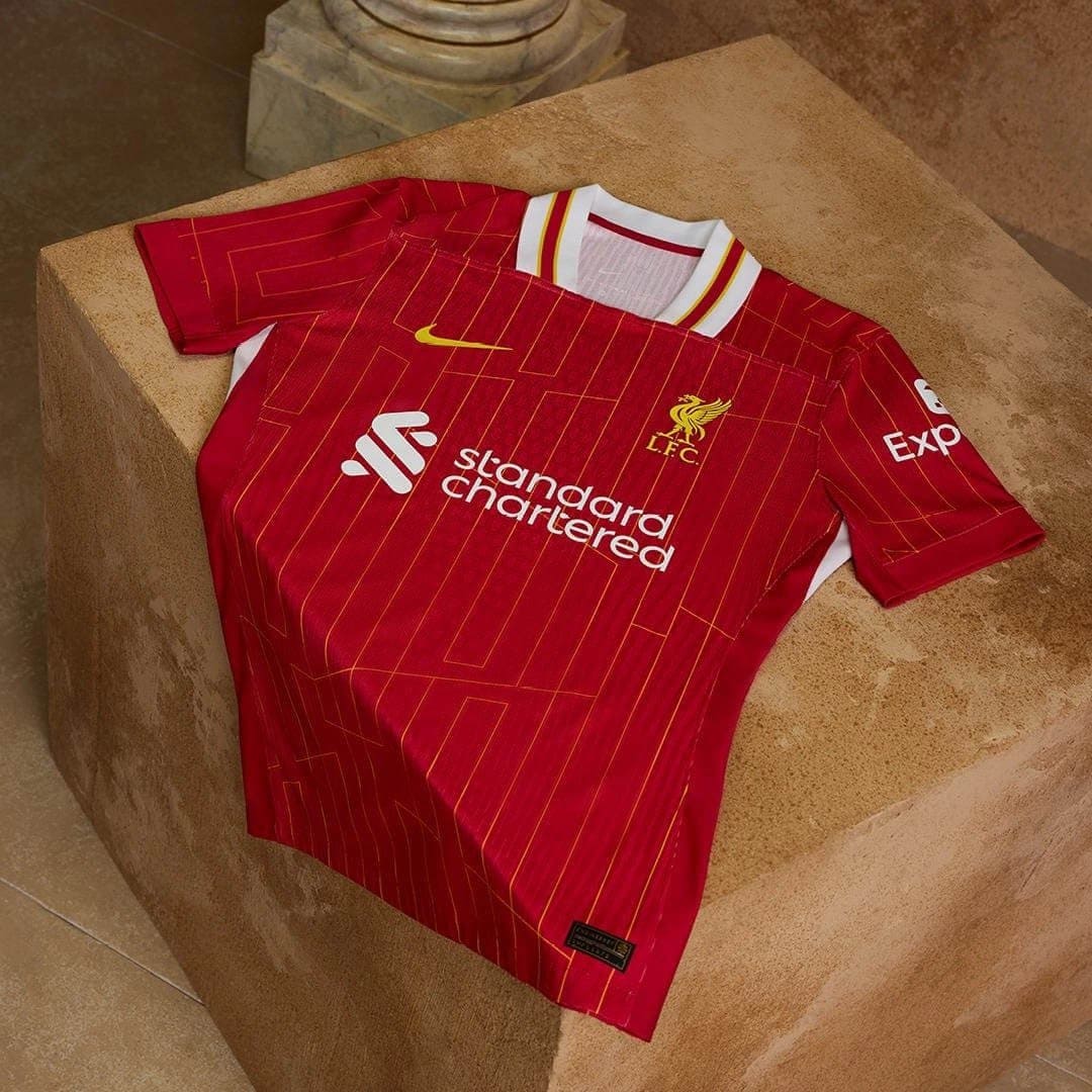 rn-image_picker_lib_temp_4e8a24b9-be7b-4889-af5d-f90758776add | AE JERSEY Red Liverpool FC home jersey with yellow crest, Nike swoosh, and 'Standard Chartered' sponsor laid on a beige stone block surface.