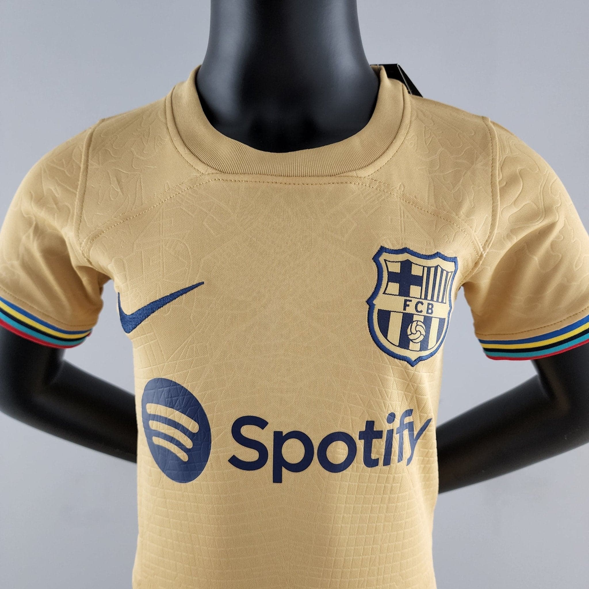 rn-image_picker_lib_temp_59875e69-ec53-4955-9b4a-be04c386f2e6 | AE JERSEY Close-up of a beige sports jersey on a mannequin, showing a blue Nike swoosh and a crest with FCB on the chest, plus striped sleeve edges.