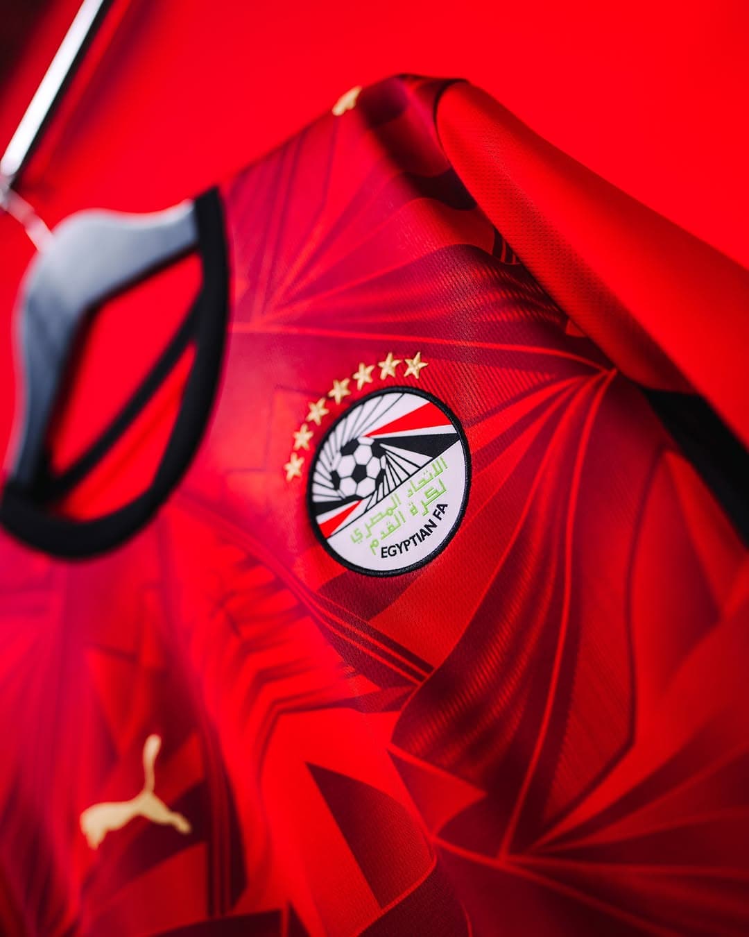 rn-image_picker_lib_temp_6c7cbc60-7a77-4fe6-b659-5a0f94e42e13 | AE JERSEY Red football jersey close-up showing the Egyptian Football Association crest with flag motif and five gold stars on the chest; Puma logo visible nearby.