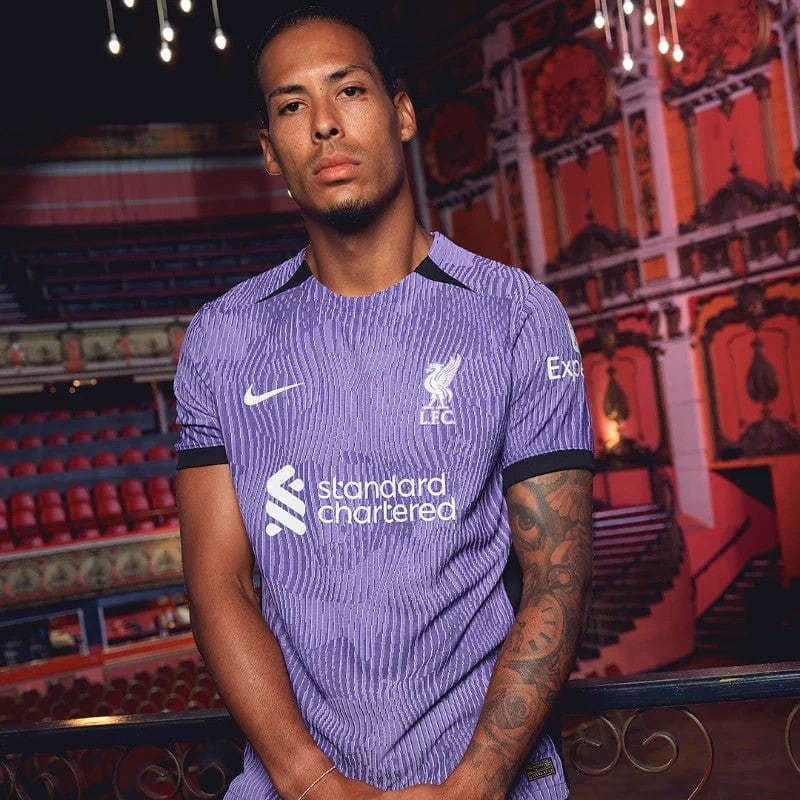 rn-image_picker_lib_temp_6f53b52f-b11c-47bb-964d-48d043efbcd1 | AE JERSEY Man wearing a purple Liverpool FC jersey posing in a theater with red seats and decorative lighting in the background.