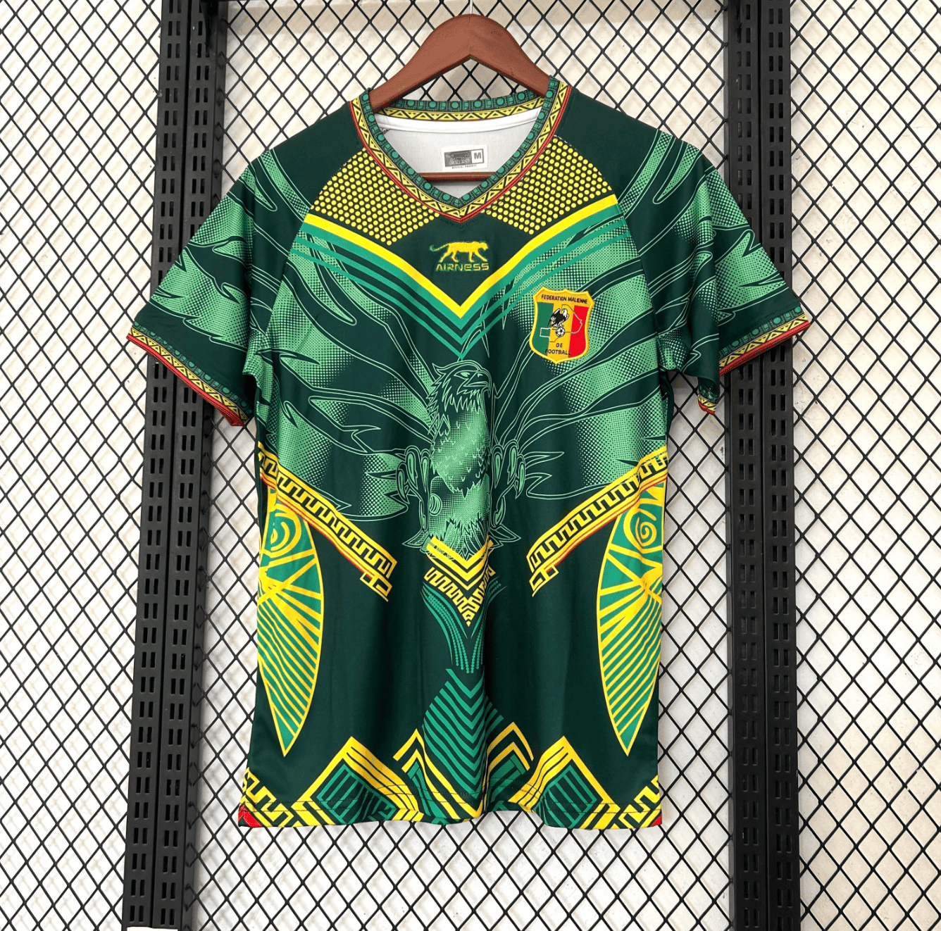 rn-image_picker_lib_temp_70bb4cd8-9534-4262-b281-61687fbd6e11 | AE JERSEY Green football jersey with yellow geometric patterns and an eagle emblem, Mali federation crest on the chest, hanging on a wooden hanger against a mesh background.