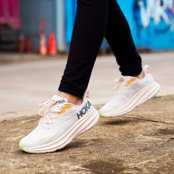 Close-up of white HOKA running shoes stepping on a sidewalk, with black pants and a colorful graffiti backdrop.