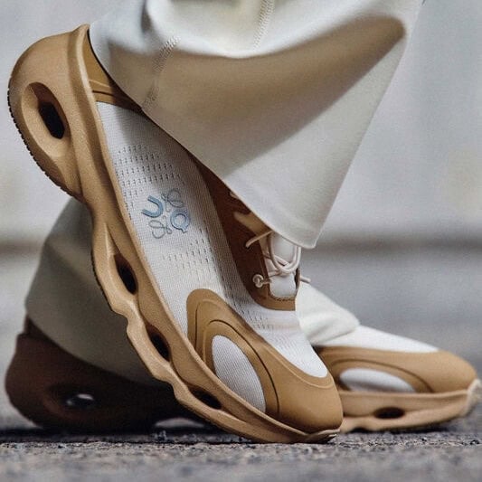Close-up of a tan-and-white athletic sneaker stepping on pavement, highlighting the thick, sculpted sole with cutouts.