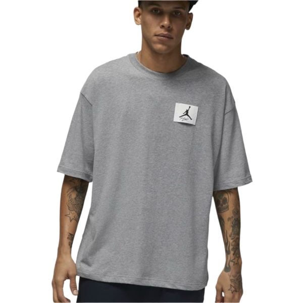 Man wearing a light gray oversized T-shirt with a small square logo on the chest, showing tattoos on both forearms.