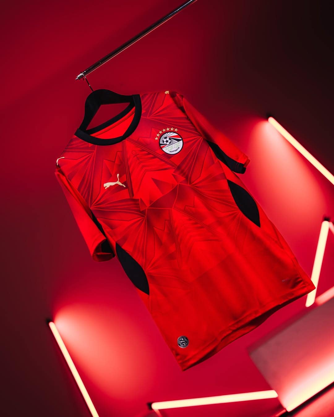 rn-image_picker_lib_temp_9477f159-7330-4ce4-b7b8-50a7b8d566d7 | AE JERSEY Red geometric-patterned football jersey with black trim, Puma logo, and club crest, hanging on a hanger under red neon lights on a dark background.